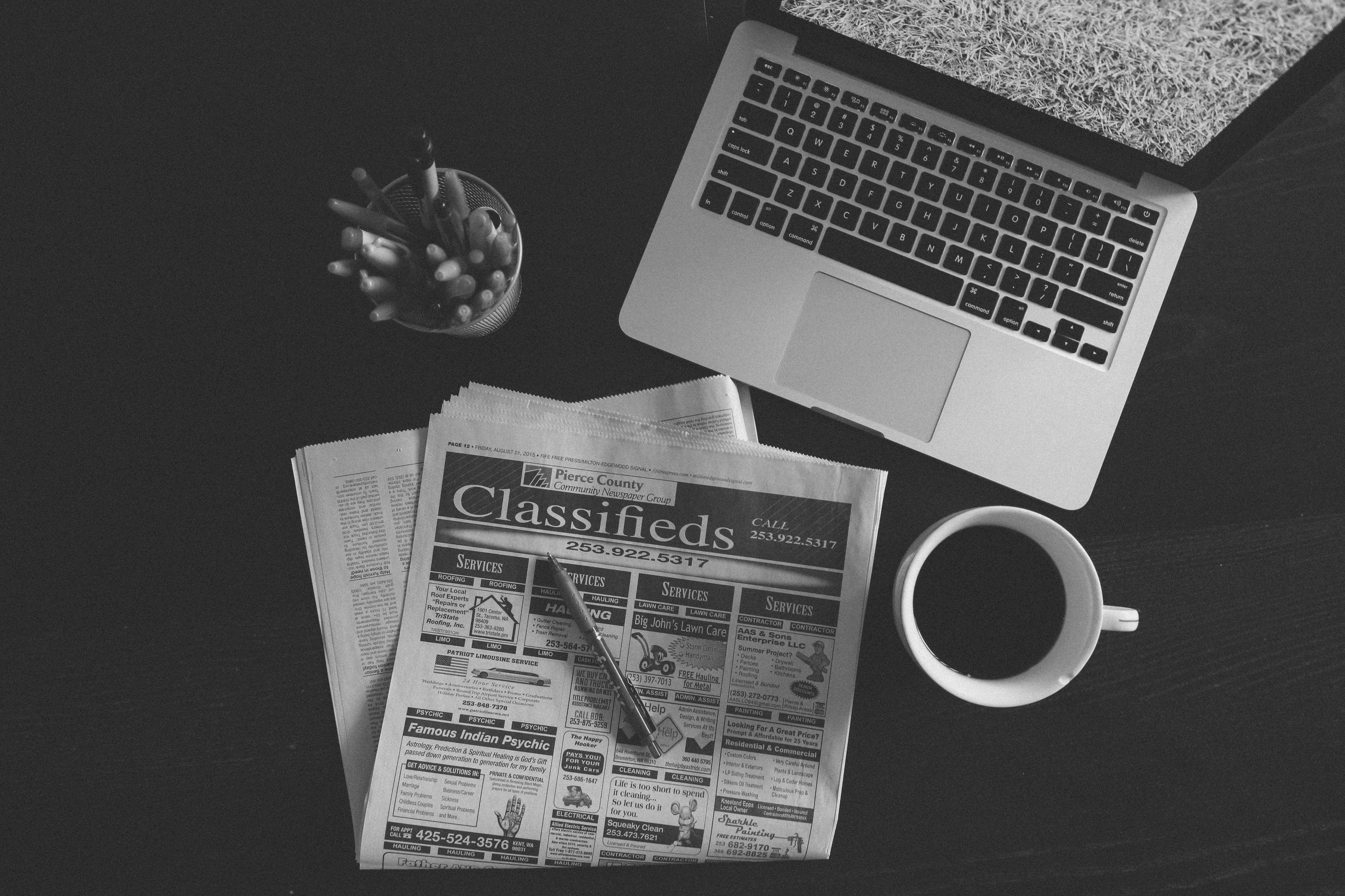 A black and white photo of an open laptop and the classifieds section of a newspaper